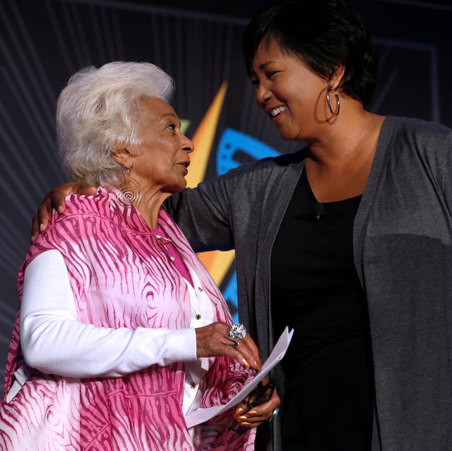 nichelle nichols and mae c jemison look at one another while standing together and speaking, nichols wears a pink vest over a white long sleeve sweater, jemison has one arm around nichols shoulders and wears a gray sweater and a black top